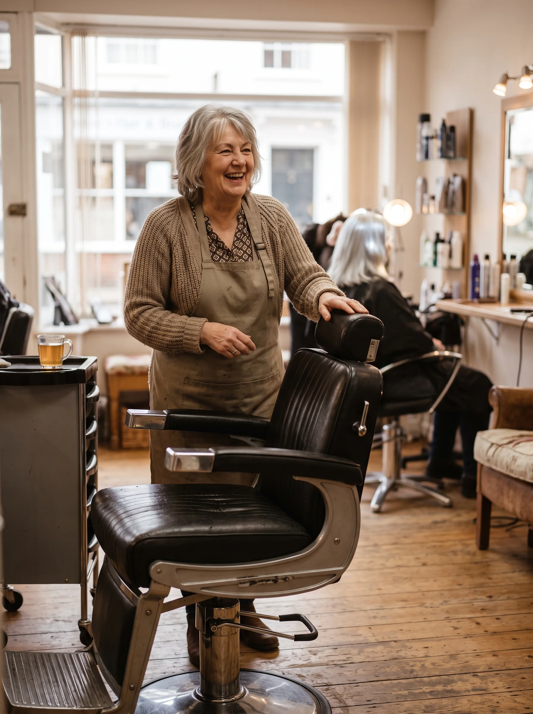 A smiling woman in a hair salon stands next to a barber chair.